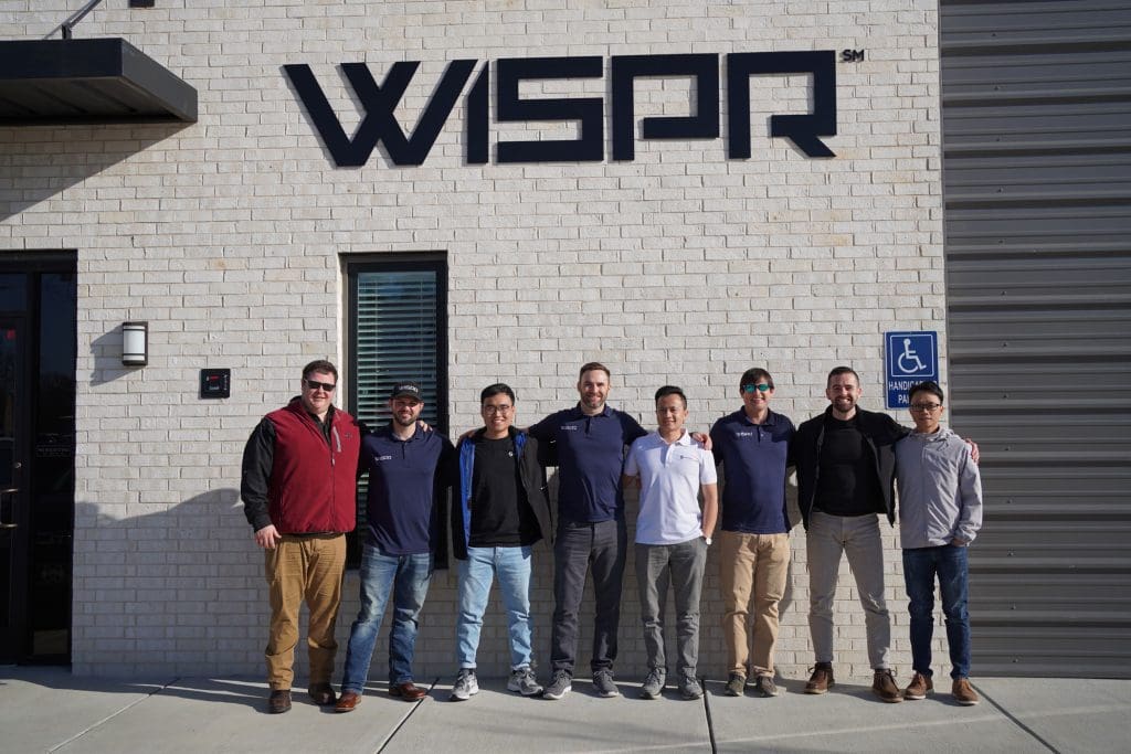 Eight men stand before a building, its sign heralding "American Made Drone by WISPR".